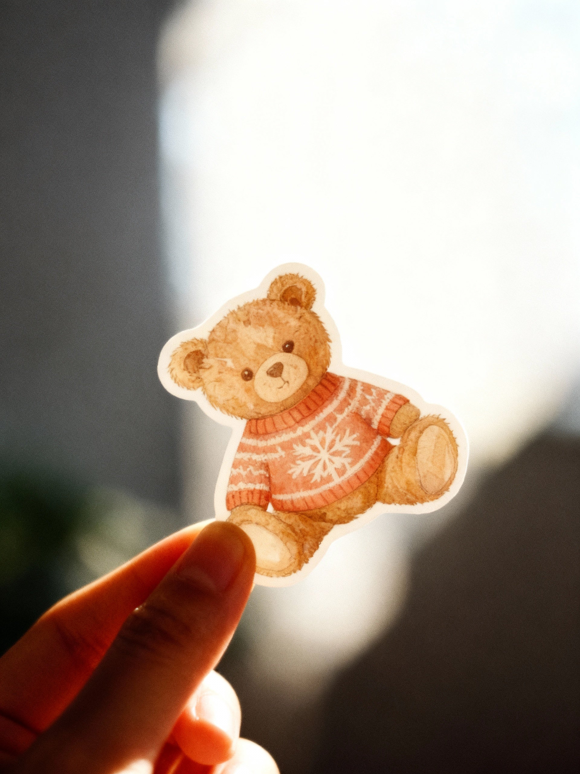 Hand holding a sticker of a teddy bear wearing a sweater with a blurred background